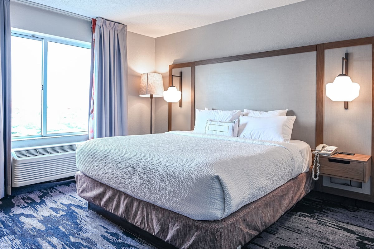 Ein Hotelzimmer mit einem großen Bett, einem Nachttisch mit Lampe, einem Telefon und einem Fenster mit Vorhängen.