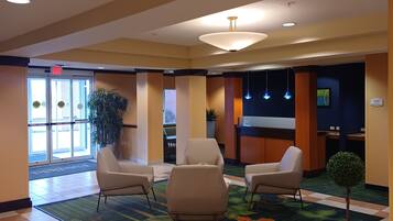 Un hall de hotel con mostrador de recepción, dos sillones, una mesita con una planta y una alfombra colorida.