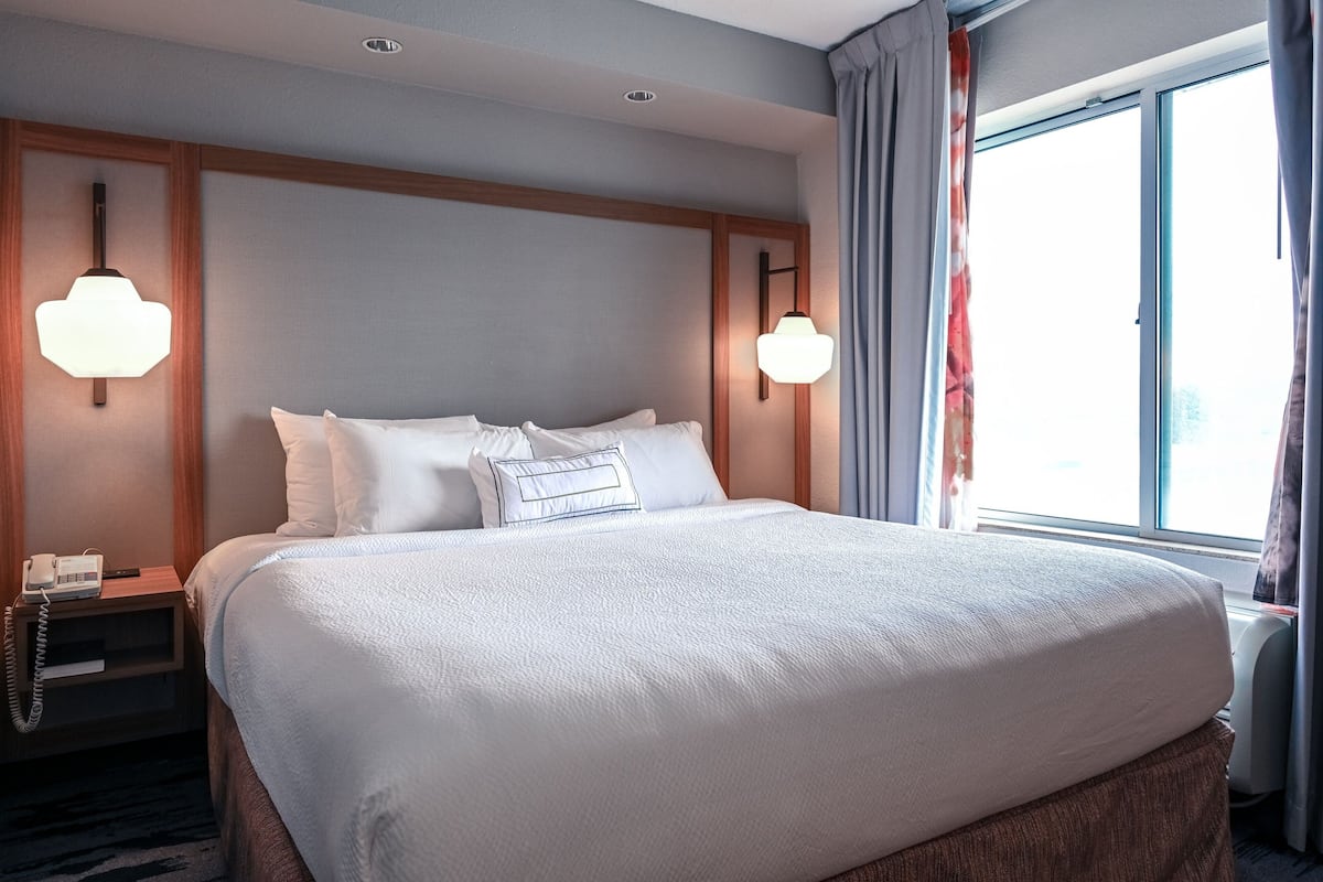 Ein Hotelzimmer mit einem großen Bett, zwei Nachttischlampen, einem Nachttisch mit Telefon und einem Fenster mit Vorhängen.