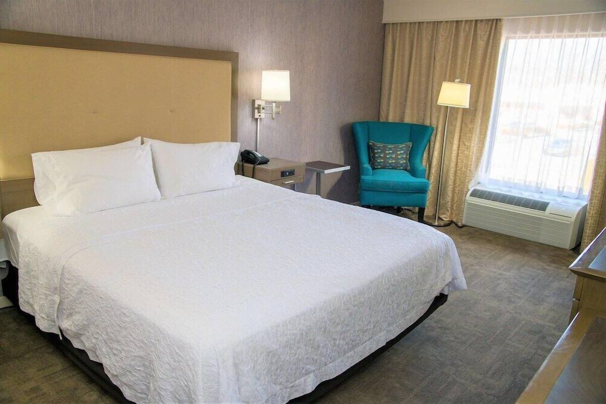 Una habitación de hotel con una cama grande, una mesita de noche con lámpara, un sillón color turquesa y una ventana con cortinas.