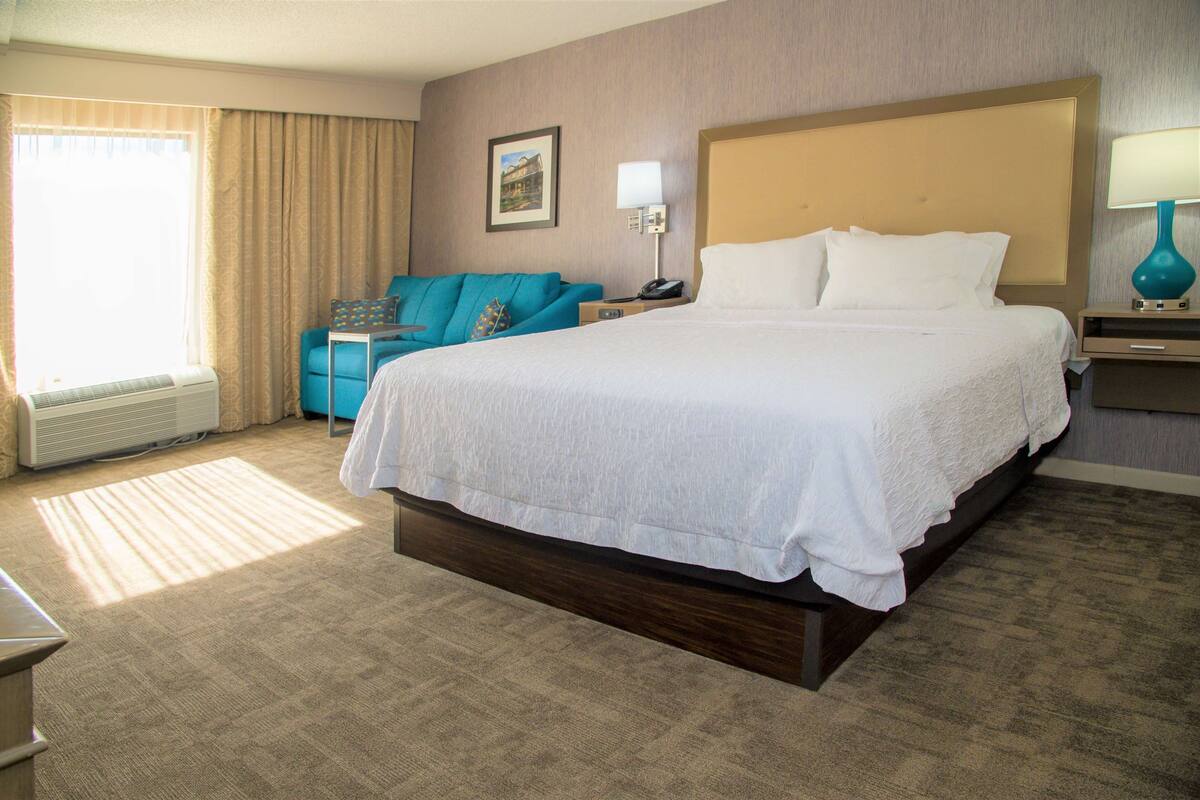 Habitación de hotel con una cama grande, un escritorio, una silla, una lámpara y una ventana con cortinas.