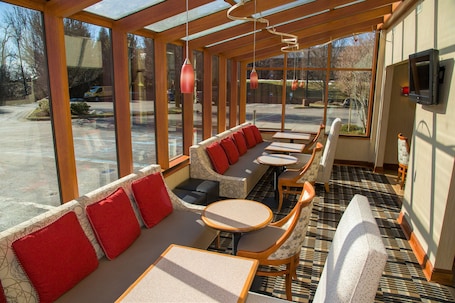 A sunlit indoor seating area with a sofa, chairs, and tables, large windows, and a television on the wall.