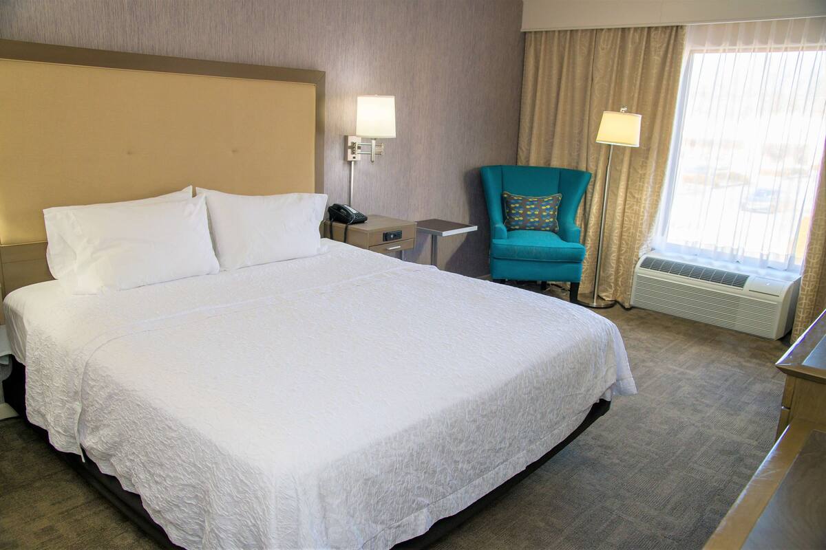 Habitación de hotel con una cama grande, un escritorio, una silla y un ventanal con cortinas.