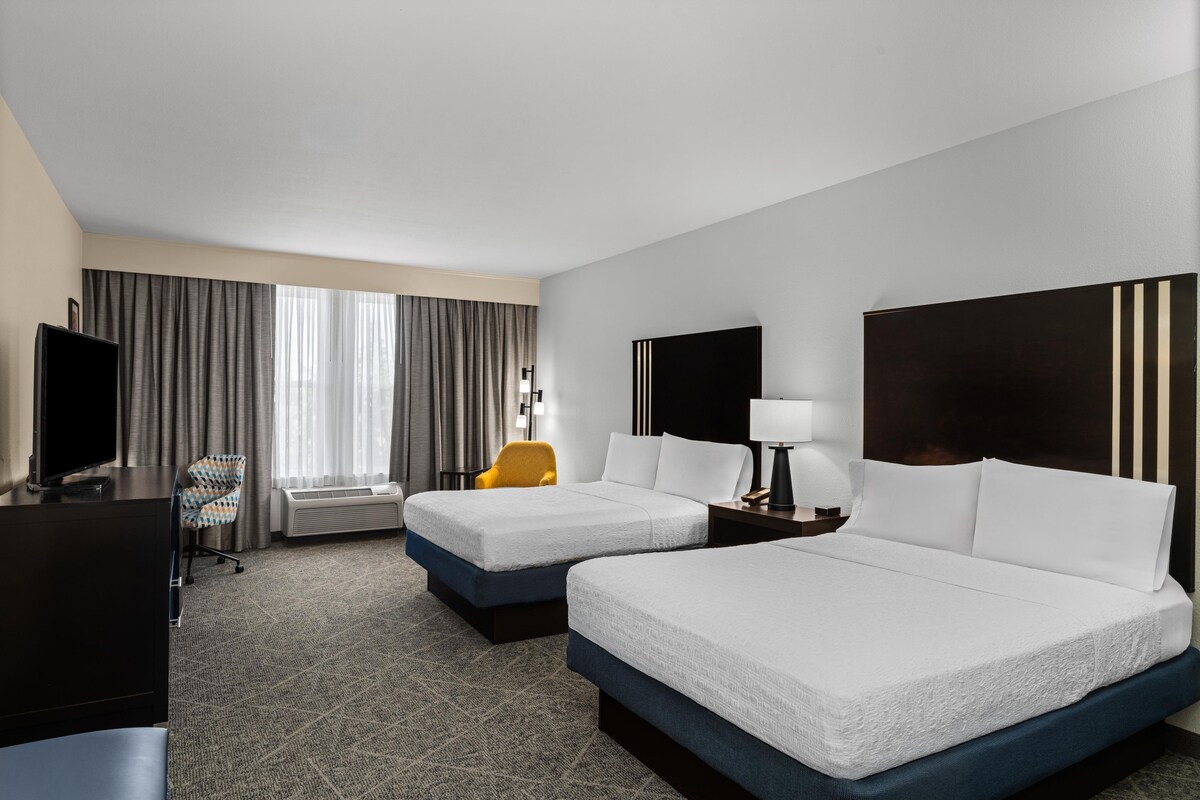 A hotel room with two beds, a desk, a chair, a TV, and a window with curtains.