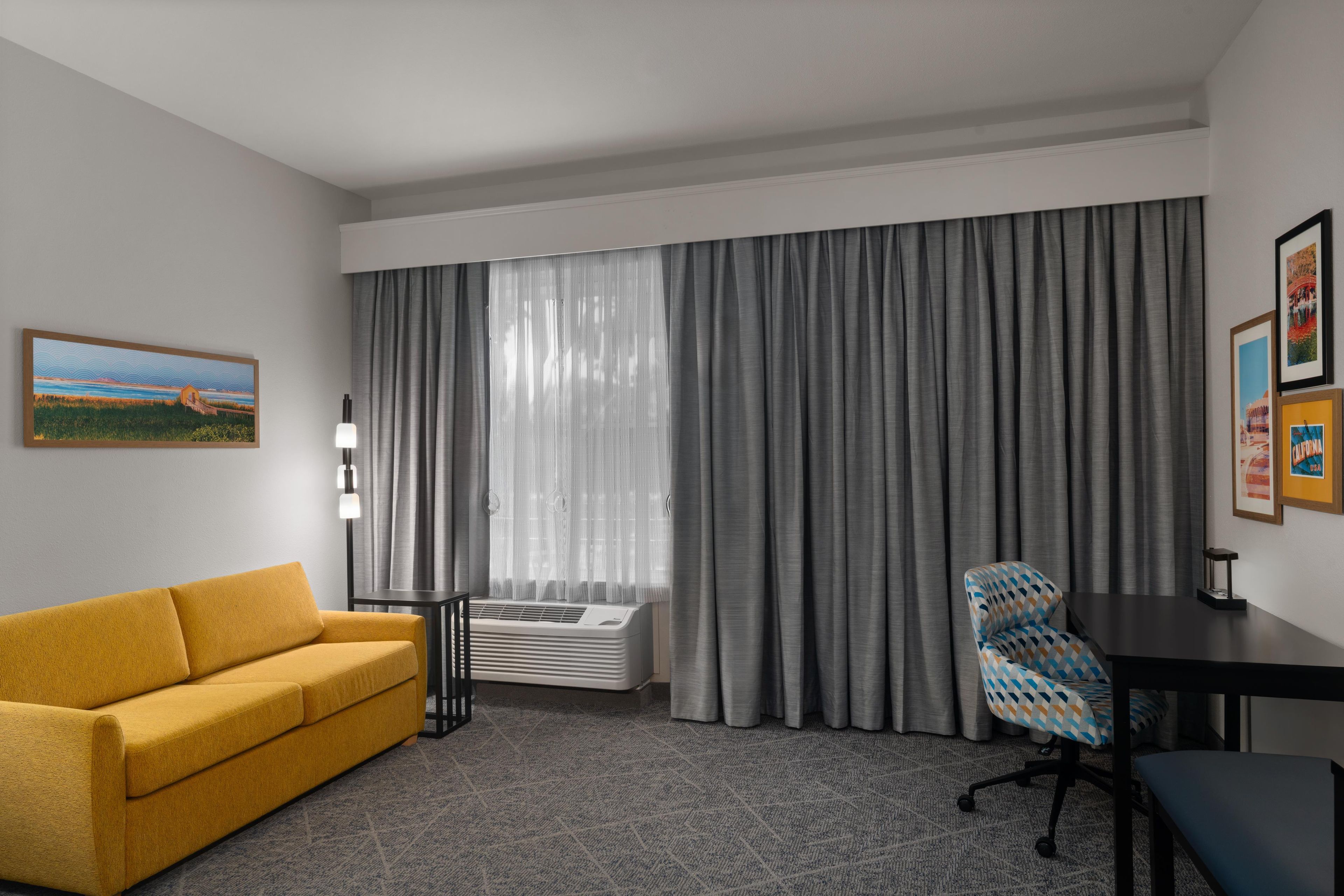 A hotel room with a sofa, a desk, a chair, a large window with curtains, and a wall-mounted air conditioner.