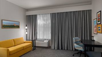 A hotel room with a sofa, a desk, a chair, a large window with curtains, and a wall-mounted air conditioner.