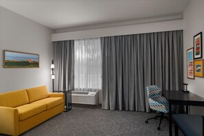 A hotel room with a sofa, a desk, a chair, a large window with curtains, and a wall-mounted air conditioner. - Hampton Inn & Suites Mountain View (Mountain View)