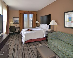 A hotel room with a large bed, two bedside tables with lamps, a television, a desk with a chair, a sofa, and a window with curtains.