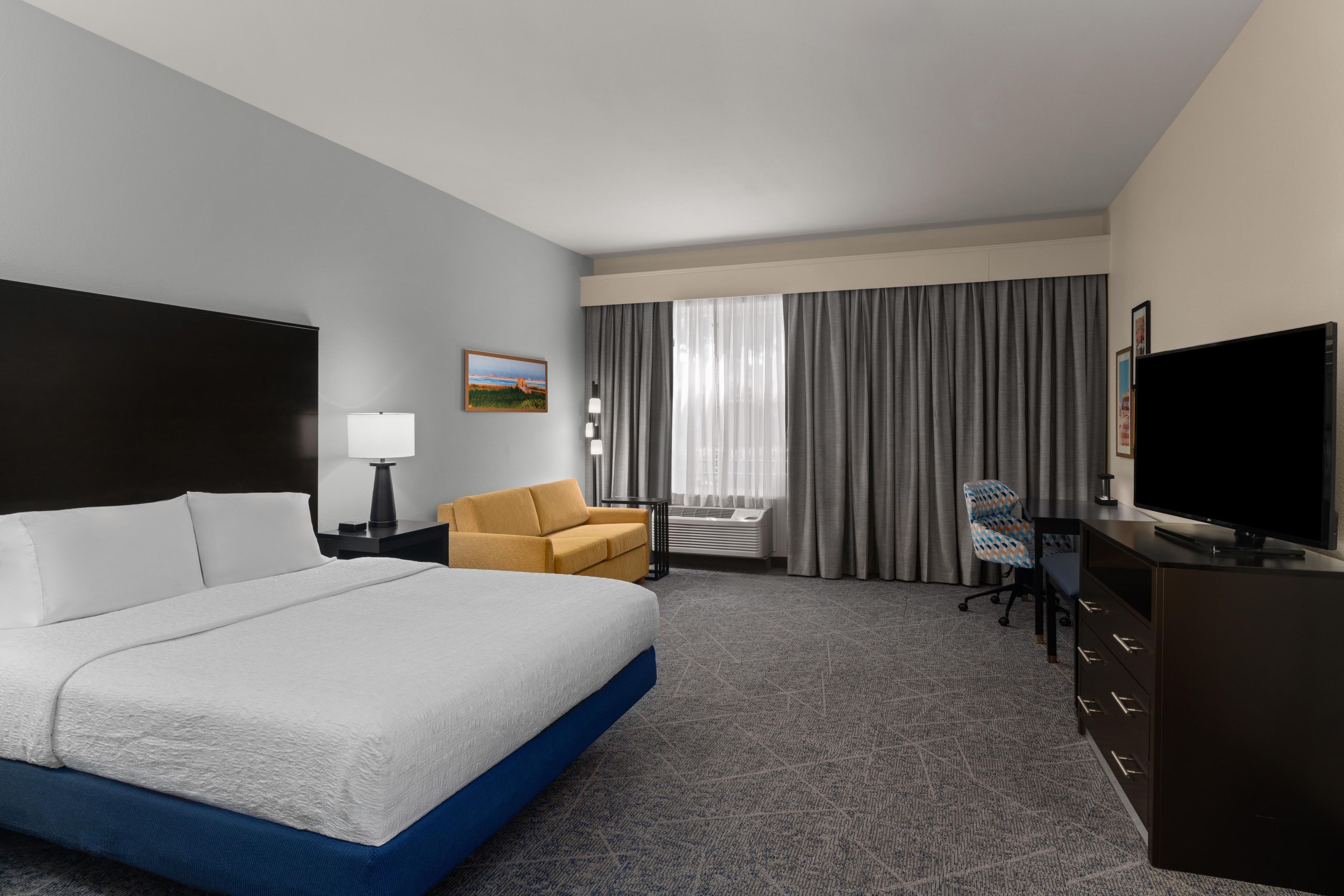 Quarto de hotel com cama grande, escrivaninha, cadeira, televisão e janela com cortinas.