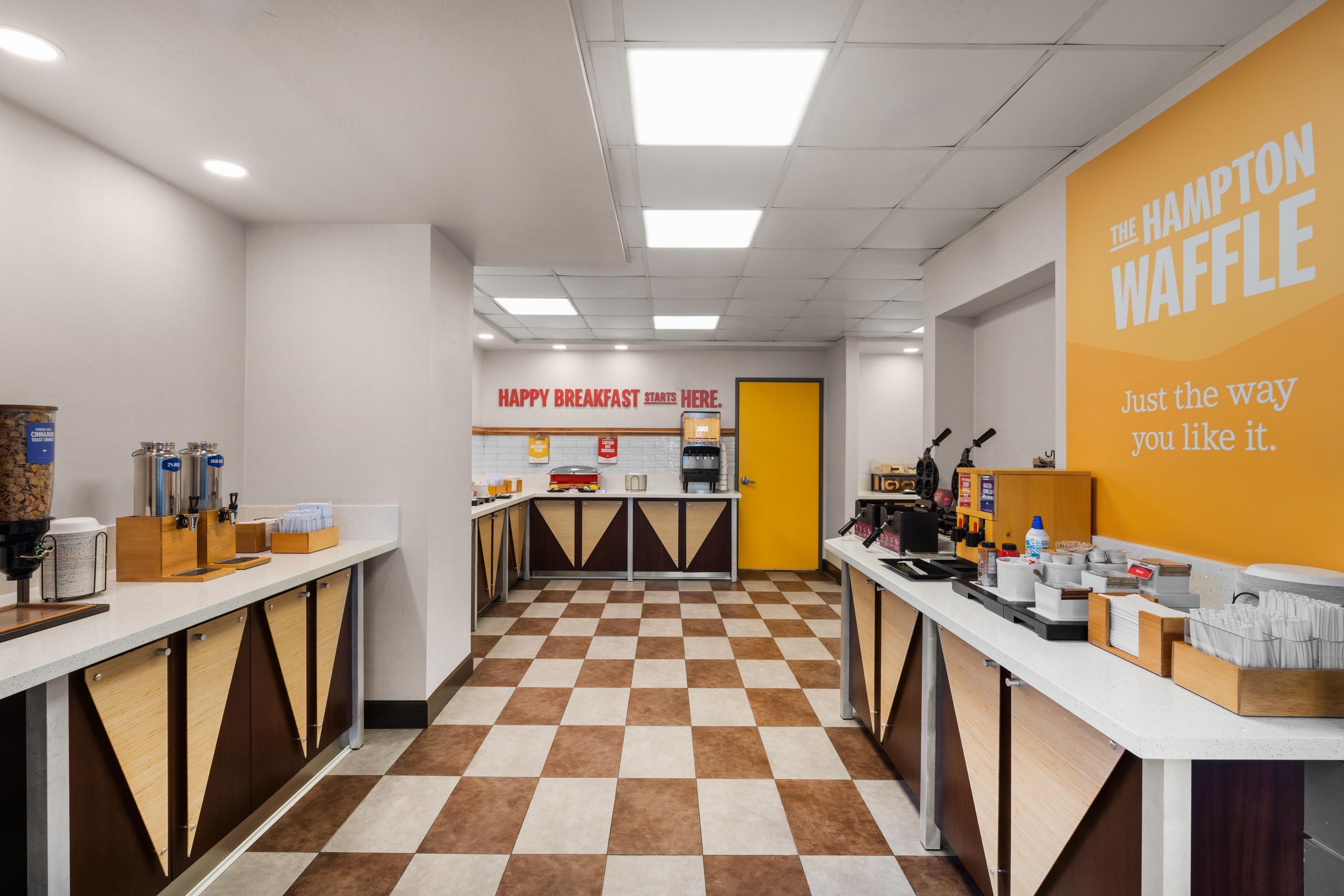 A clean, modern breakfast area with checkered flooring, a central counter, and a bright yellow wall with the Hampton Waffle logo.