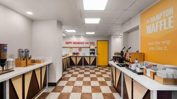 A clean, modern breakfast area with checkered flooring, a central counter, and a bright yellow wall with the Hampton Waffle logo.