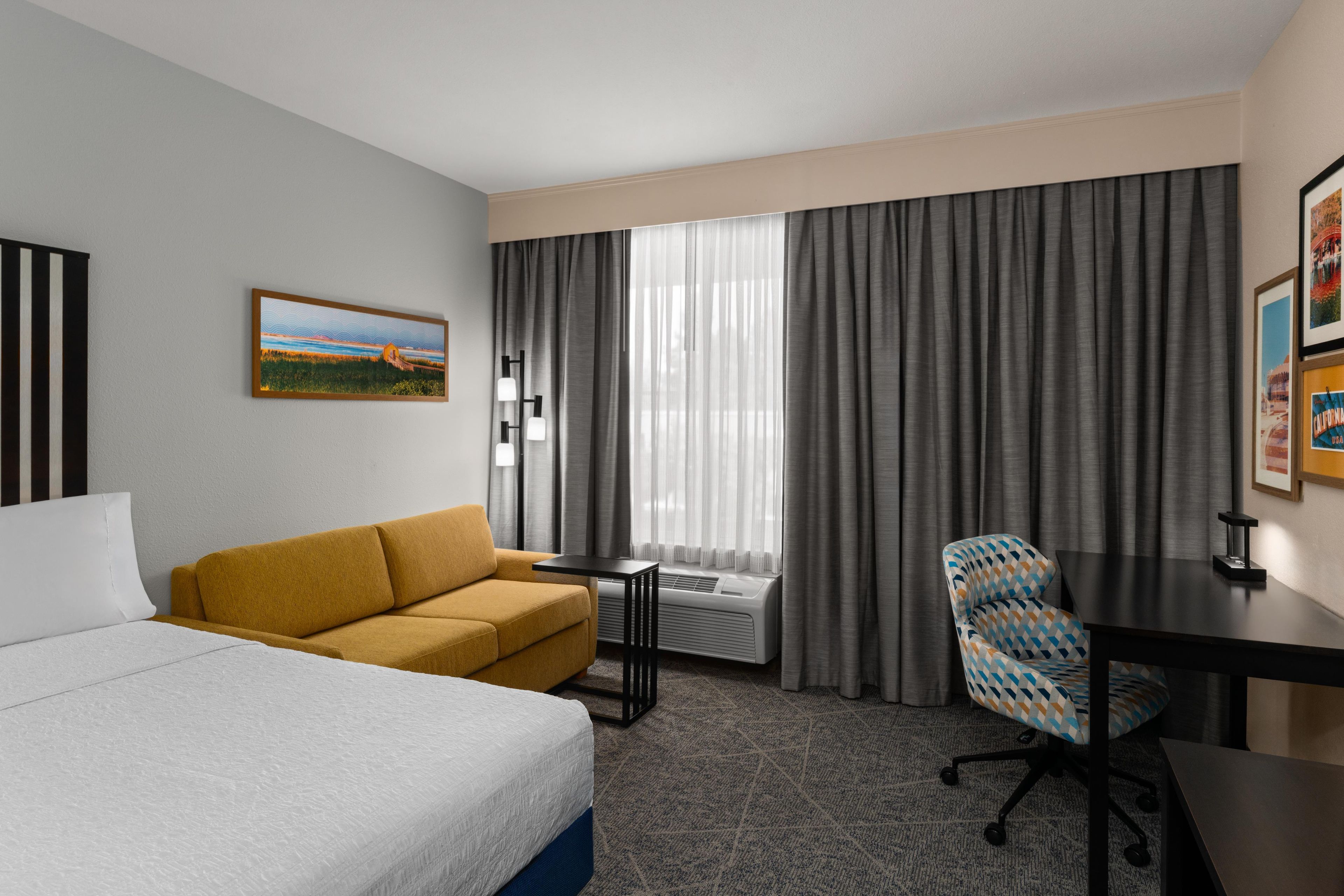 A hotel room with a bed, a sofa, a desk, a chair, a window with curtains, and a painting on the wall.