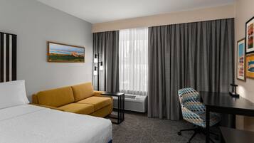 A hotel room with a bed, a sofa, a desk, a chair, a window with curtains, and a painting on the wall.
