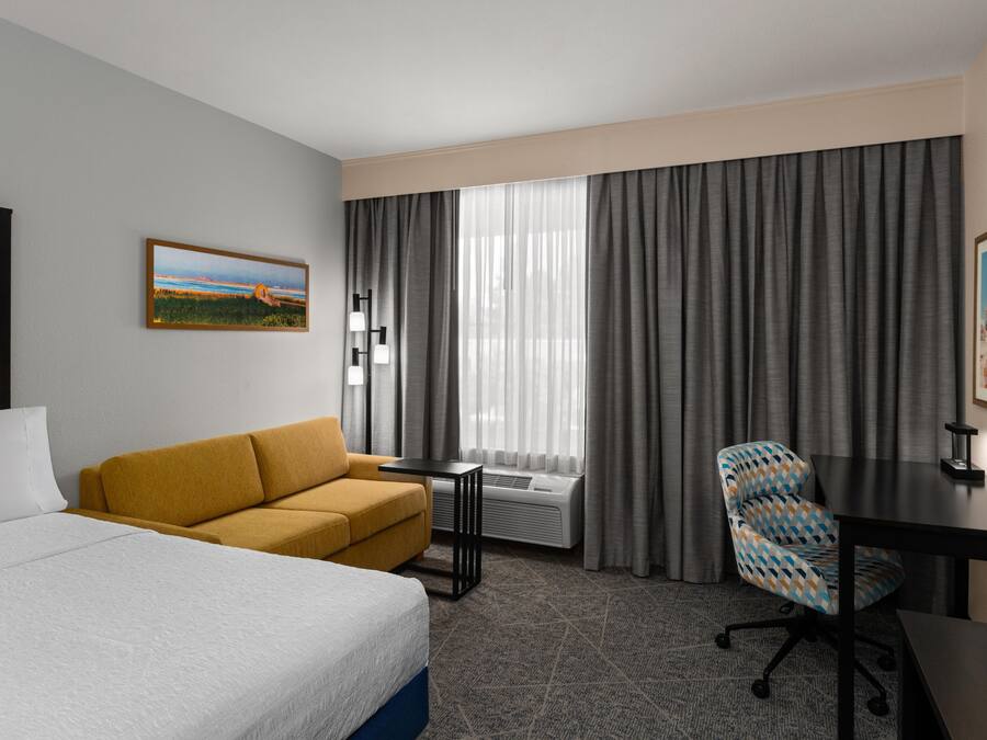 A hotel room with a bed, a sofa, a desk, a chair, a window with curtains, and a painting on the wall.