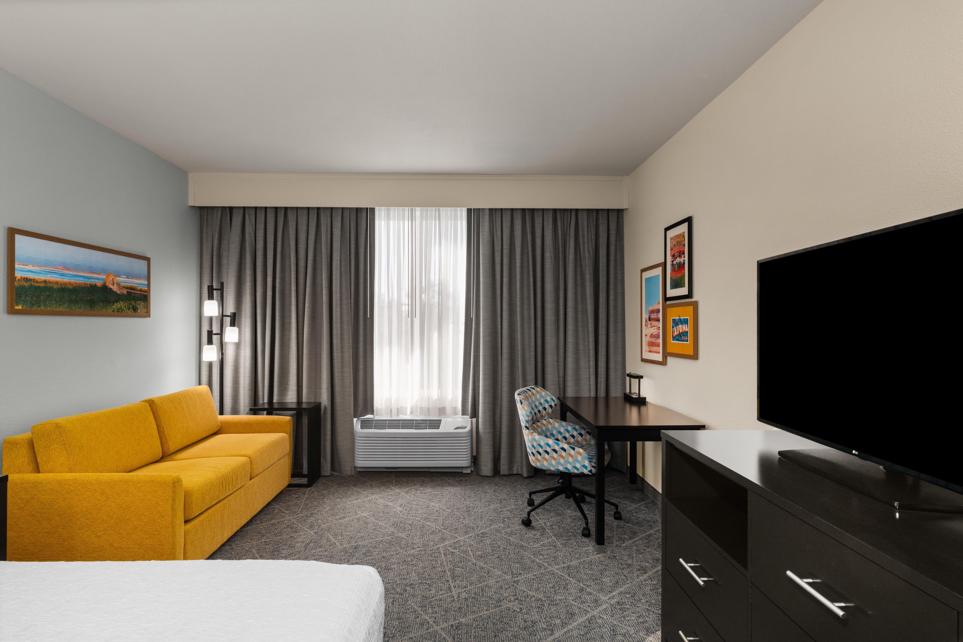 A hotel room with a bed, a yellow sofa, a desk with a chair, a television, and a large window with curtains.