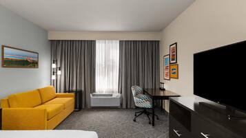 A hotel room with a bed, a yellow sofa, a desk with a chair, a television, and a large window with curtains.
