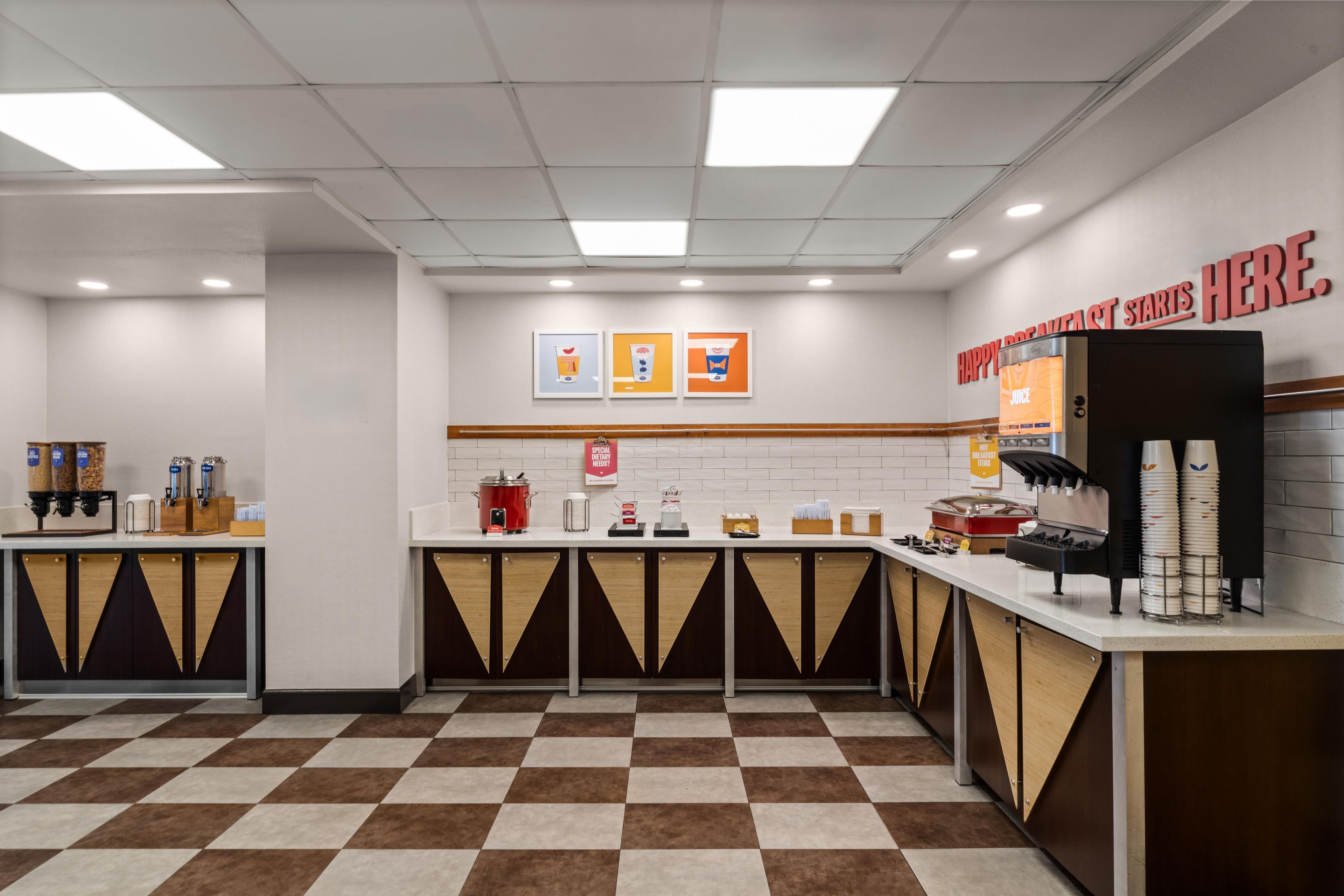 Un espace réservé au petit-déjeuner, avec un sol à carreaux, une machine à café, un distributeur de jus de fruits et un panneau indiquant l’heure du début du service.