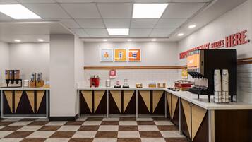 Un espace réservé au petit-déjeuner, avec un sol à carreaux, une machine à café, un distributeur de jus de fruits et un panneau indiquant l’heure du début du service.