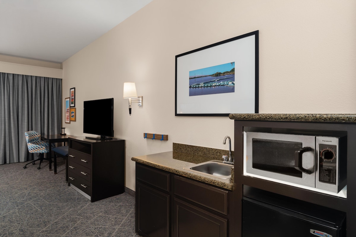 A hotel room with a flat-screen TV, a desk, a chair, a framed picture on the wall, a microwave, and a sink.