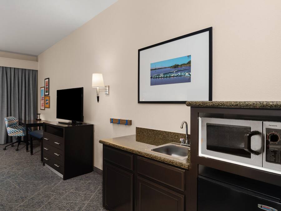 A hotel room with a flat-screen TV, a desk, a chair, a framed picture on the wall, a microwave, and a sink.