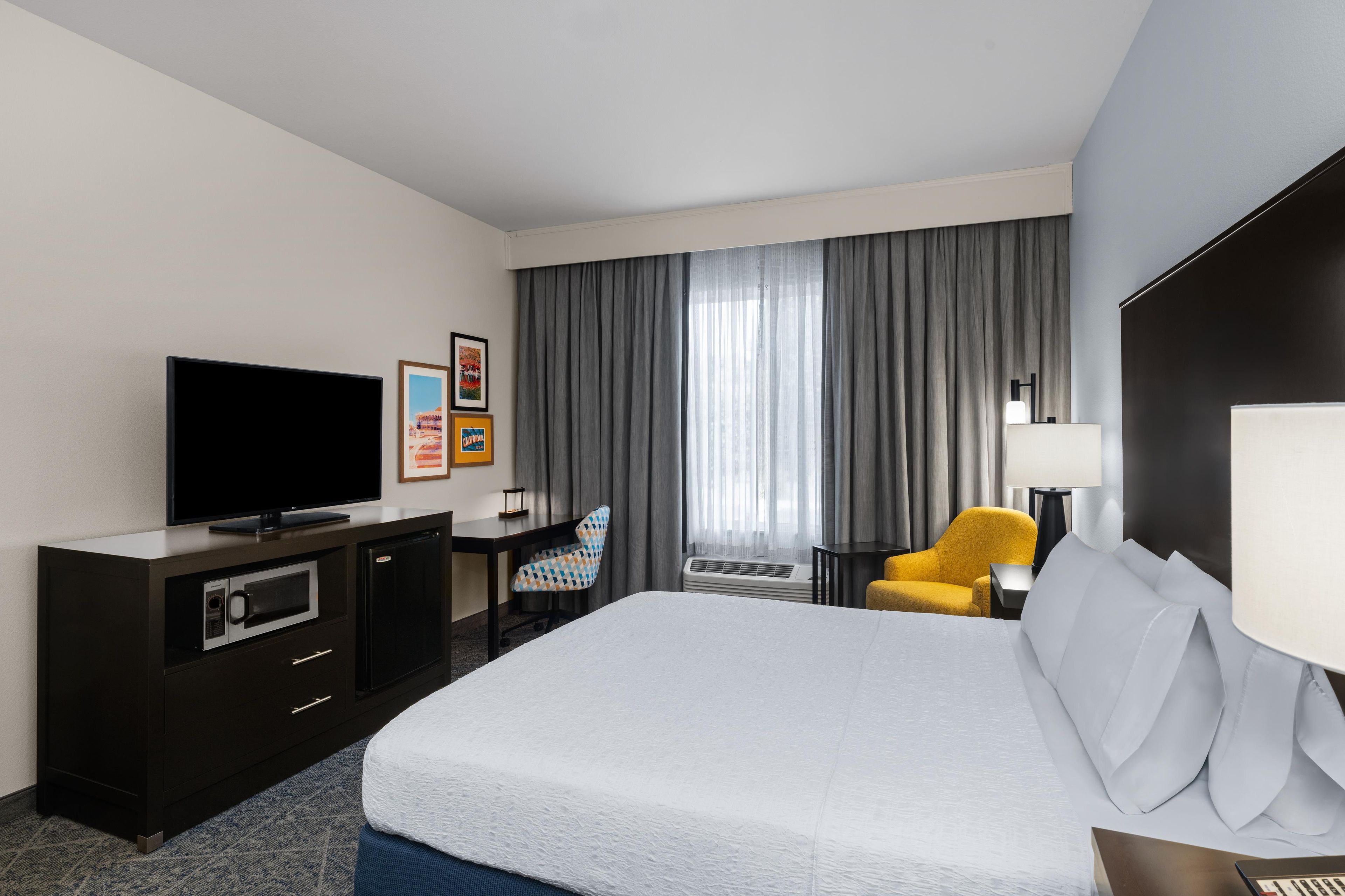 Une chambre d’hôtel avec un lit, un bureau, une télévision, une chaise et une fenêtre avec des rideaux.