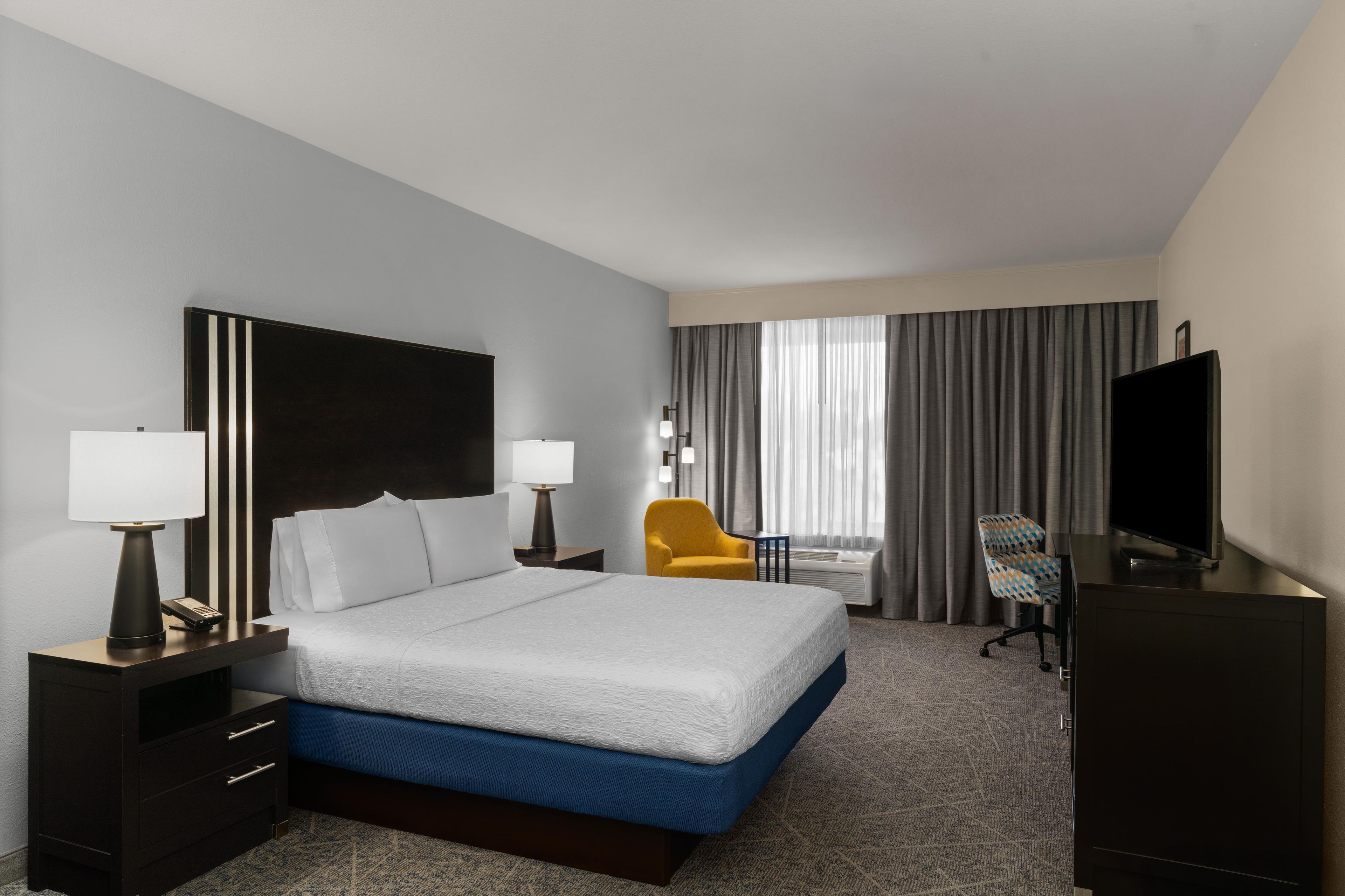 Quarto de hotel com cama, duas mesas de cabeceira com luminárias, escrivaninha com cadeira, televisão e uma janela grande com cortinas.