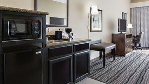 Quarto de hotel compacto com cozinha compacta, incluindo micro-ondas, cafeteira e pequena geladeira. Possui uma única cadeira e uma escrivaninha com televisão.