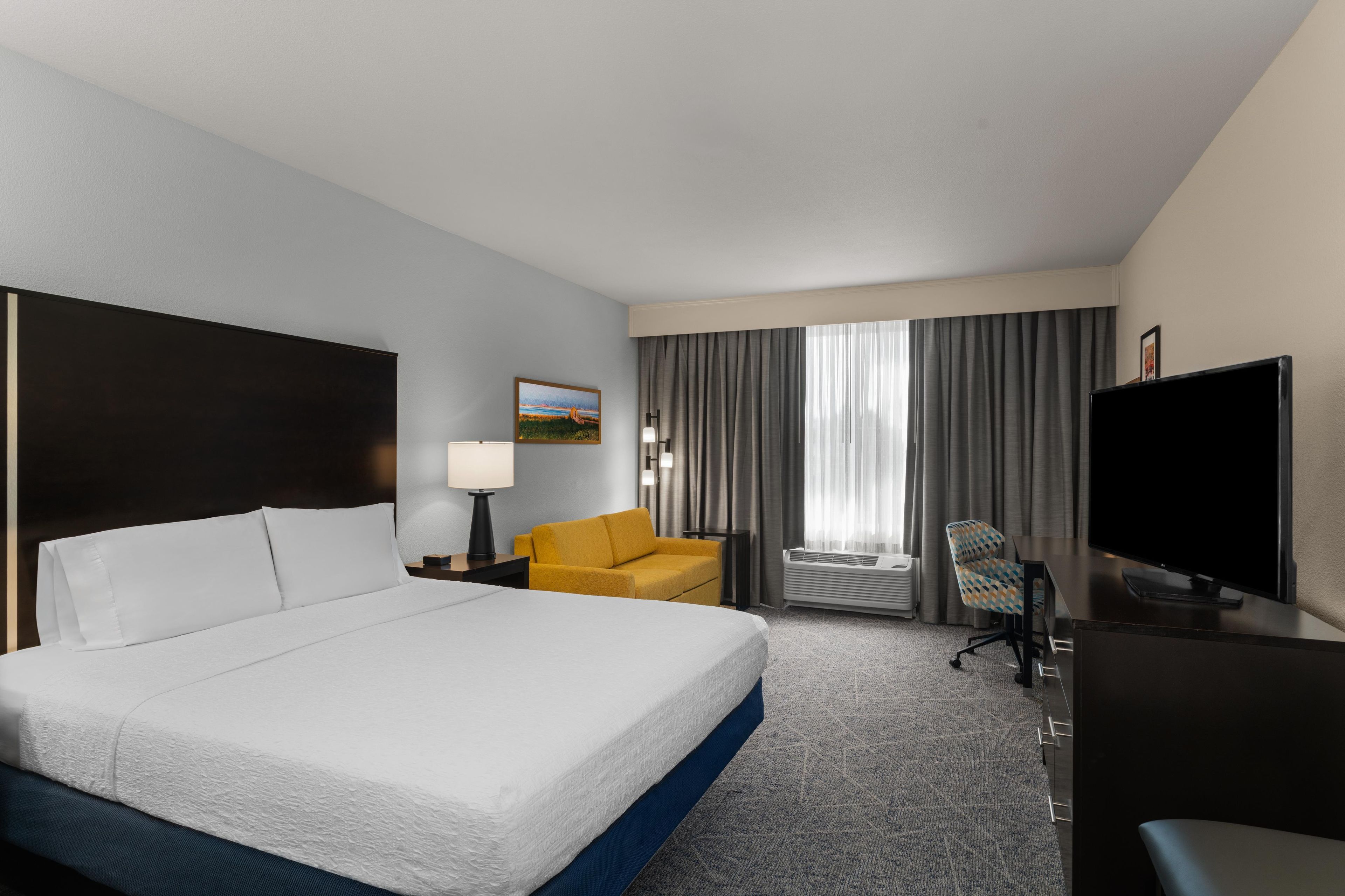 Une chambre d’hôtel avec un grand lit, un bureau doté d’un téléviseur à écran plat, un fauteuil jaune et une fenêtre avec des rideaux.