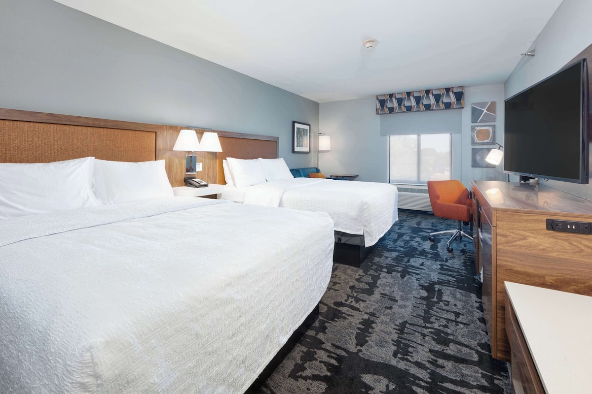 A hotel room with two beds, a desk, a chair, a TV, and a window.