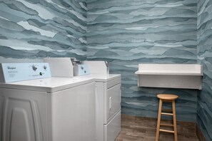 A laundry room with a front-loading washer and dryer, a wooden stool, and a wall-mounted shelf.
