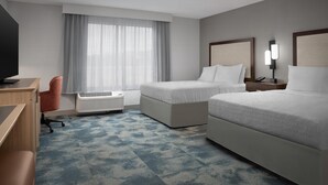 A hotel room with two beds, a desk, a chair, a TV, a window with curtains, and a wall-mounted lamp.