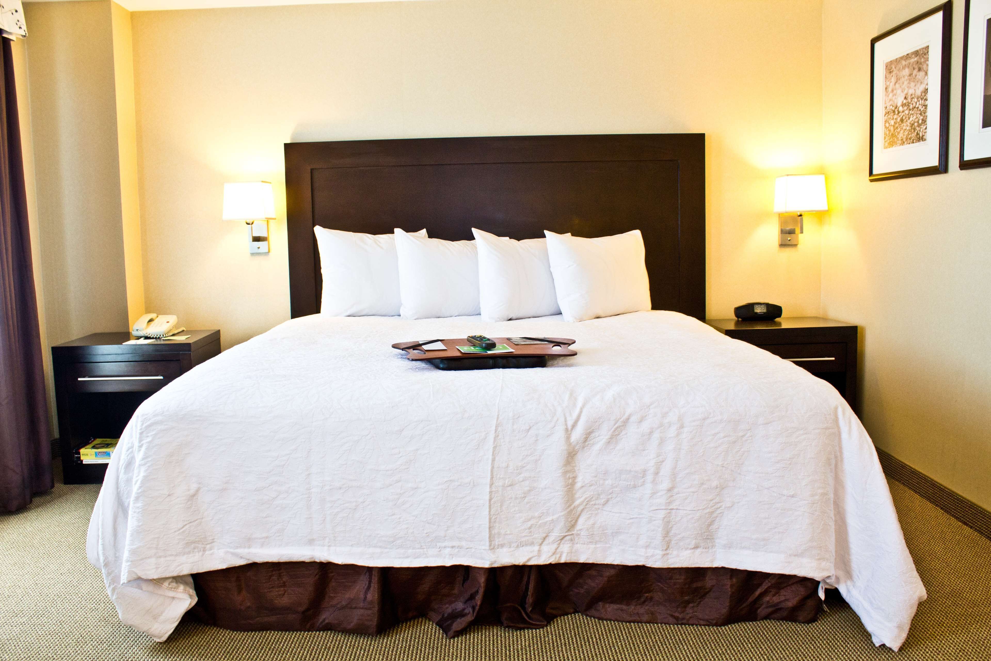 Quarto de hotel com uma cama grande, duas mesas de cabeceira, um criado-mudo, uma luminária e um quadro emoldurado na parede.