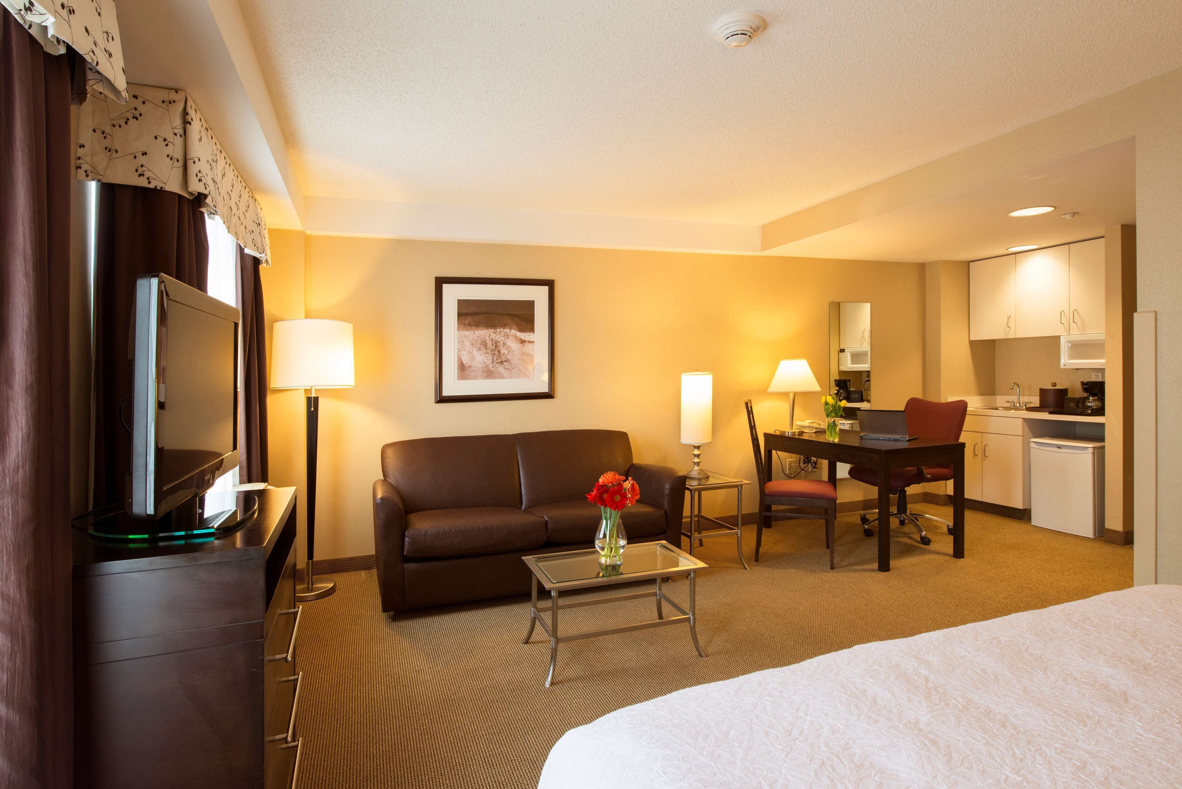 A hotel room with a bed, a desk with a chair, a sofa, a TV, a lamp, and a kitchenette.