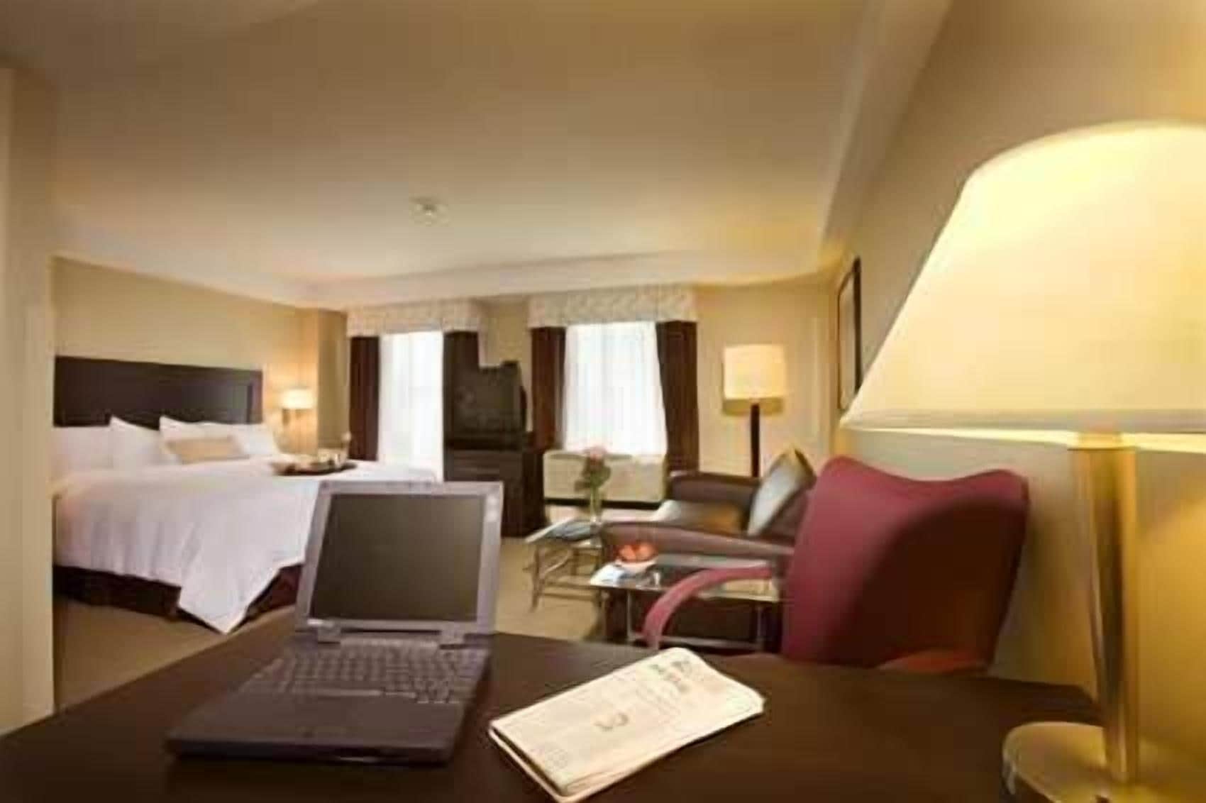 A hotel room with a bed, a desk with a laptop, a chair, and a lamp.