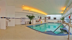 Una piscina interior amb sòl de rajola, una escala de piscina i algunes cadires i plantes al voltant de la zona de la piscina.