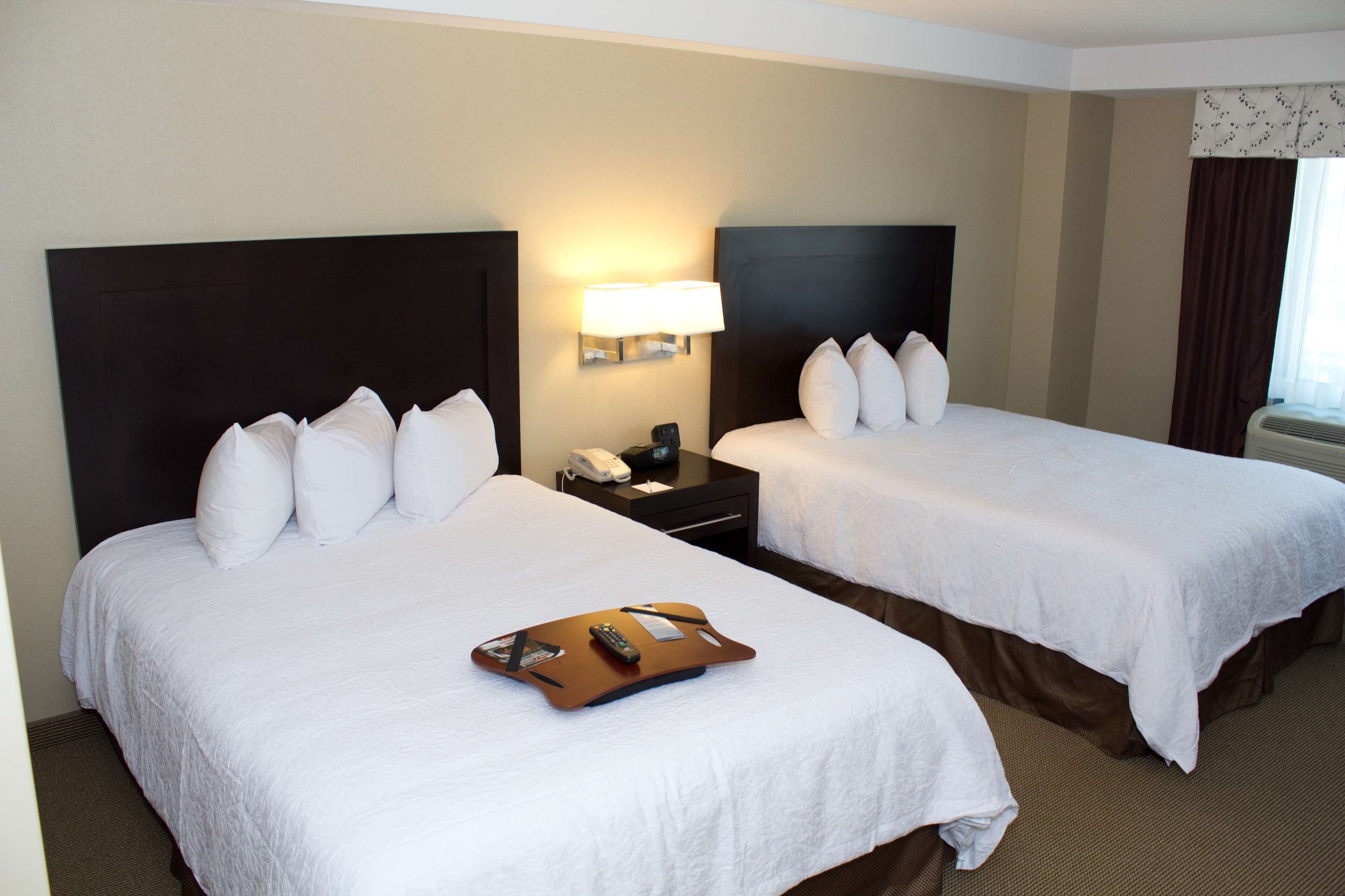 A hotel room with two beds, a bedside table with a lamp, and a tray with a remote control.