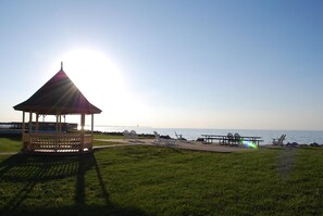 A gazebo with a sloped roof, a picnic table, and chairs in a grassy area by the water.
