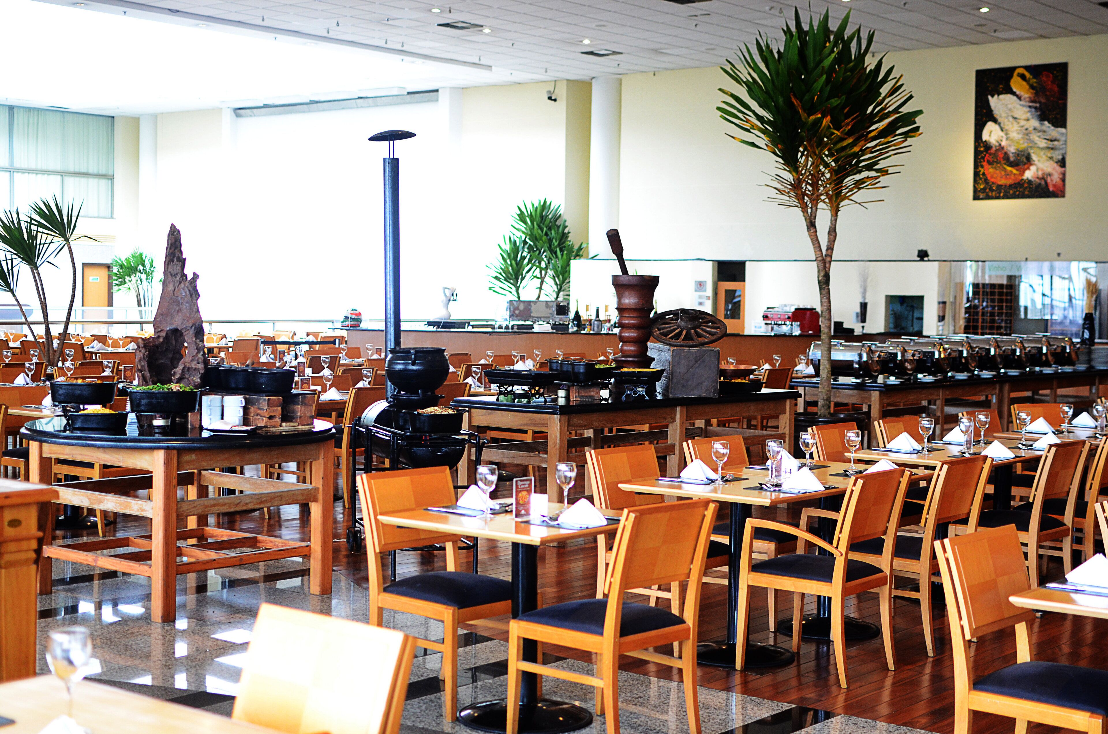 A spacious restaurant with multiple tables and chairs, a large wooden table centerpiece, and potted plants.