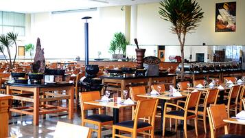 A spacious restaurant with multiple tables and chairs, a large wooden table centerpiece, and potted plants.