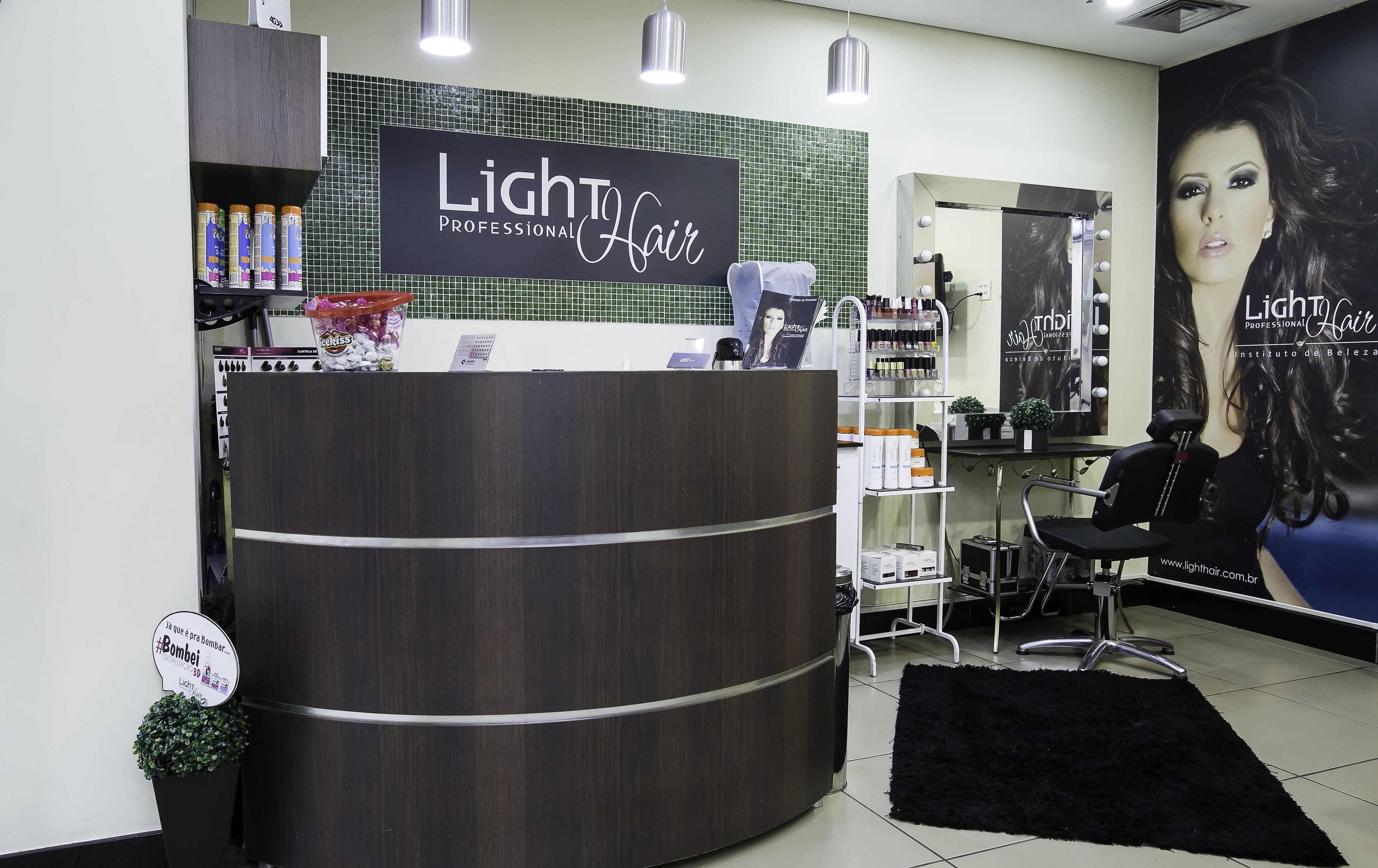 Interior de salão de beleza com balcão de recepção, produtos para cabelo, espelho com iluminação e um cartaz grande.