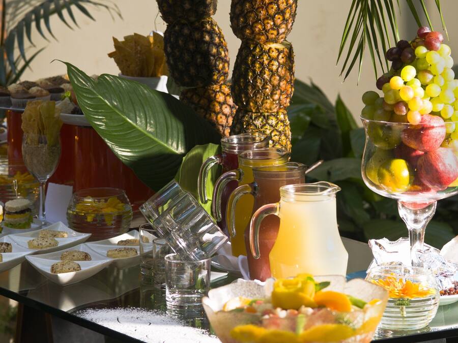 Mesa de buffet con diversos alimentos, bebidas y un fondo tropical.