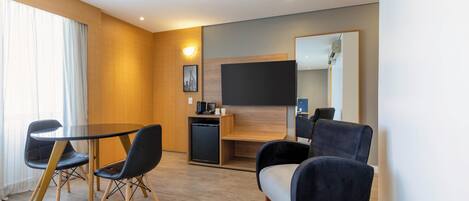 Una habitación de hotel moderna con piso de madera, una mesa de comedor redonda con sillas, una pequeña kitchenette, un televisor de pantalla plana montado en la pared y un espejo grande.