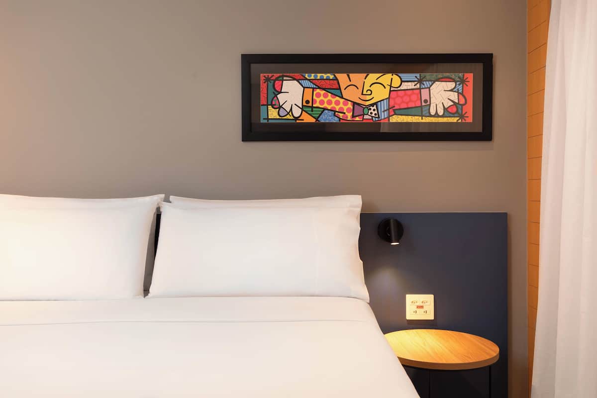 Una cama bien tendida con sábanas blancas, una obra de arte enmarcada encima y una pequeña mesa redonda de madera al lado.