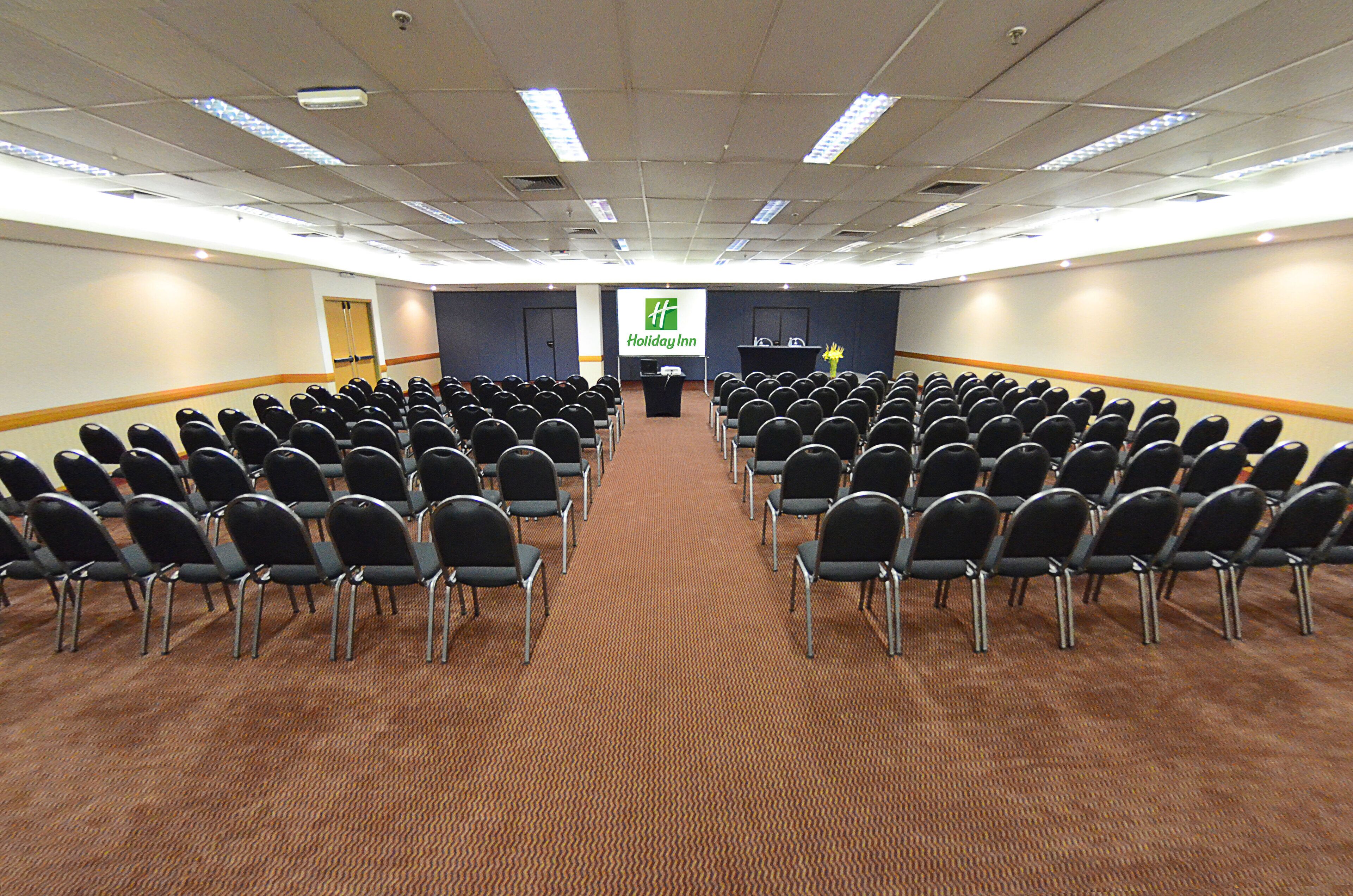A conference room with rows of chairs facing a podium, a carpeted floor, and a Holiday Inn sign on the wall.