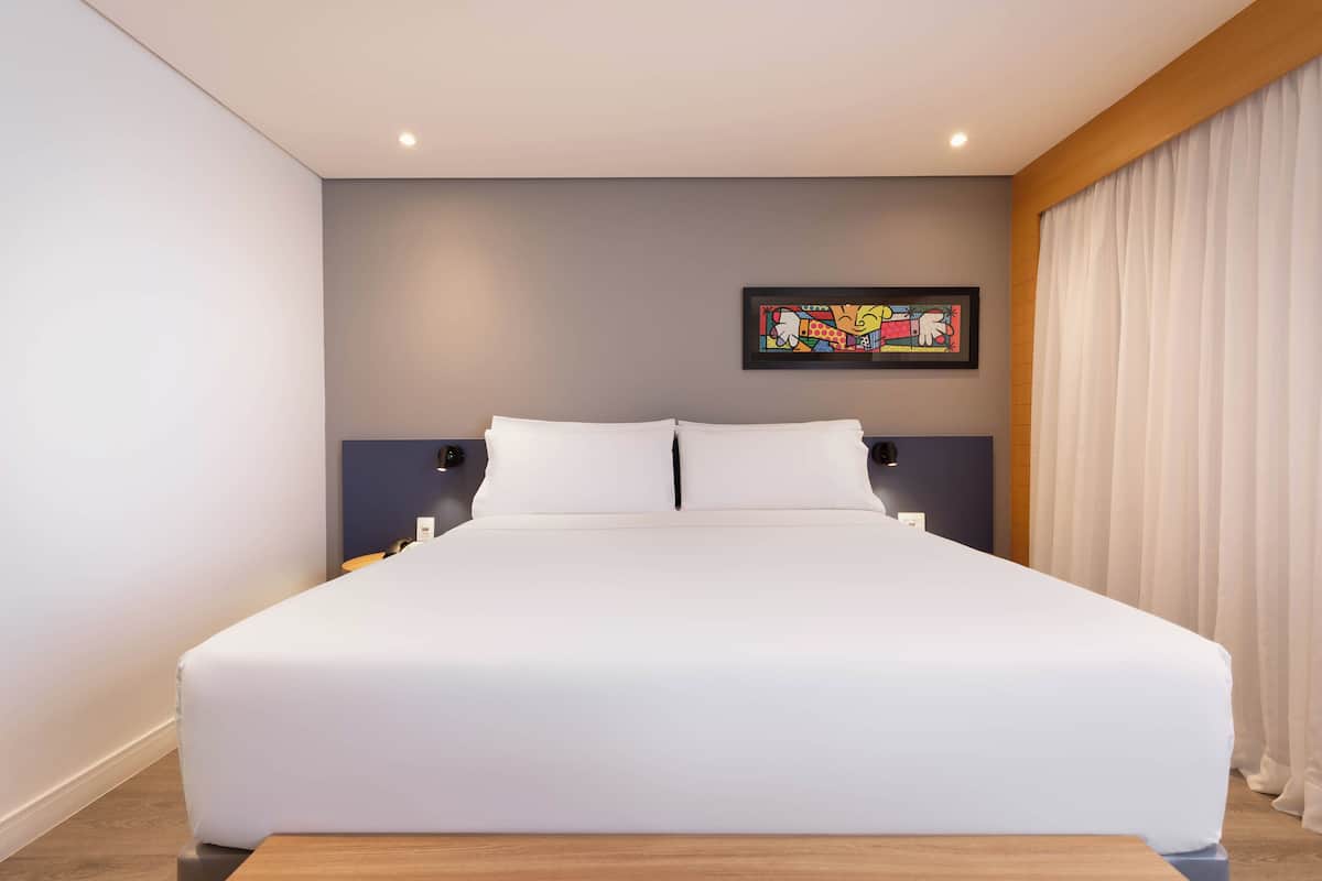Una habitación de hotel moderna con una cama grande, una mesita de noche, una obra de arte en la pared y una ventana con cortinas.