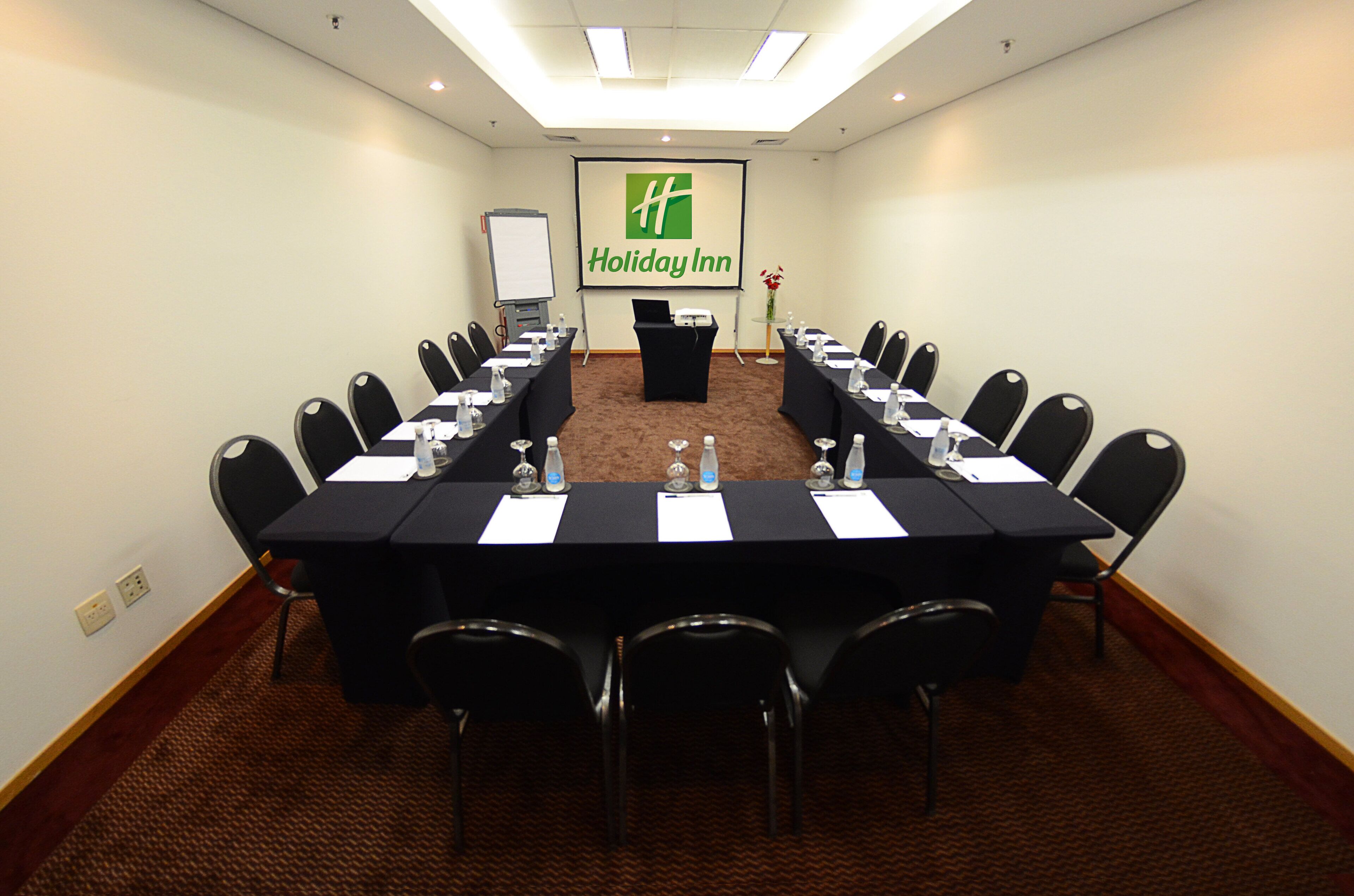 Un salón de conferencias con una larga mesa, sillas, un atril y una pantalla que muestra el logotipo de Holiday Inn.