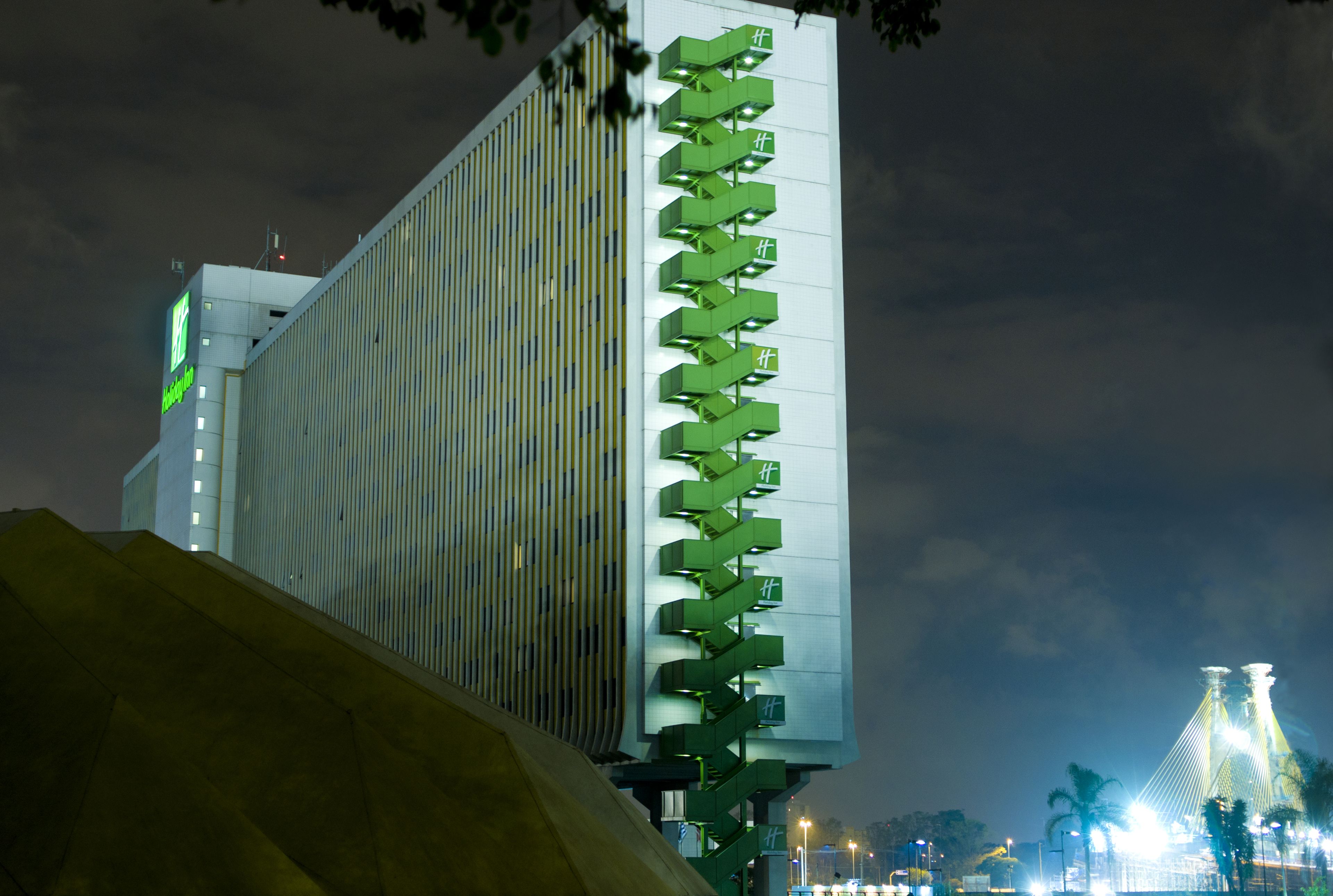 A modern building with a vertical green sign with white text, a large yellow structure in the foreground, and a bridge with lights in the background.