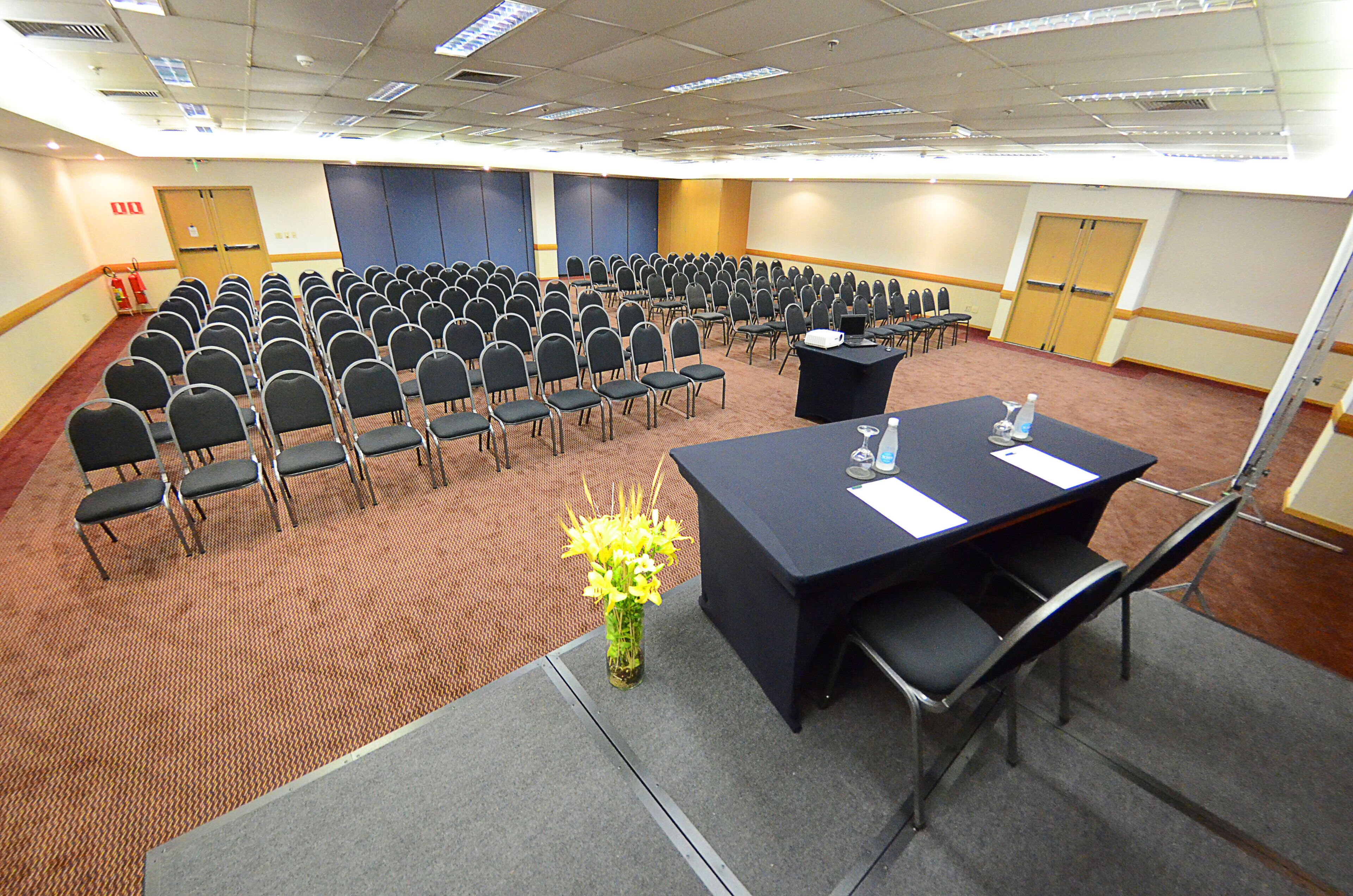 Un salón de conferencias con filas de sillas, una mesa con un jarrón de flores y una pantalla de proyector.