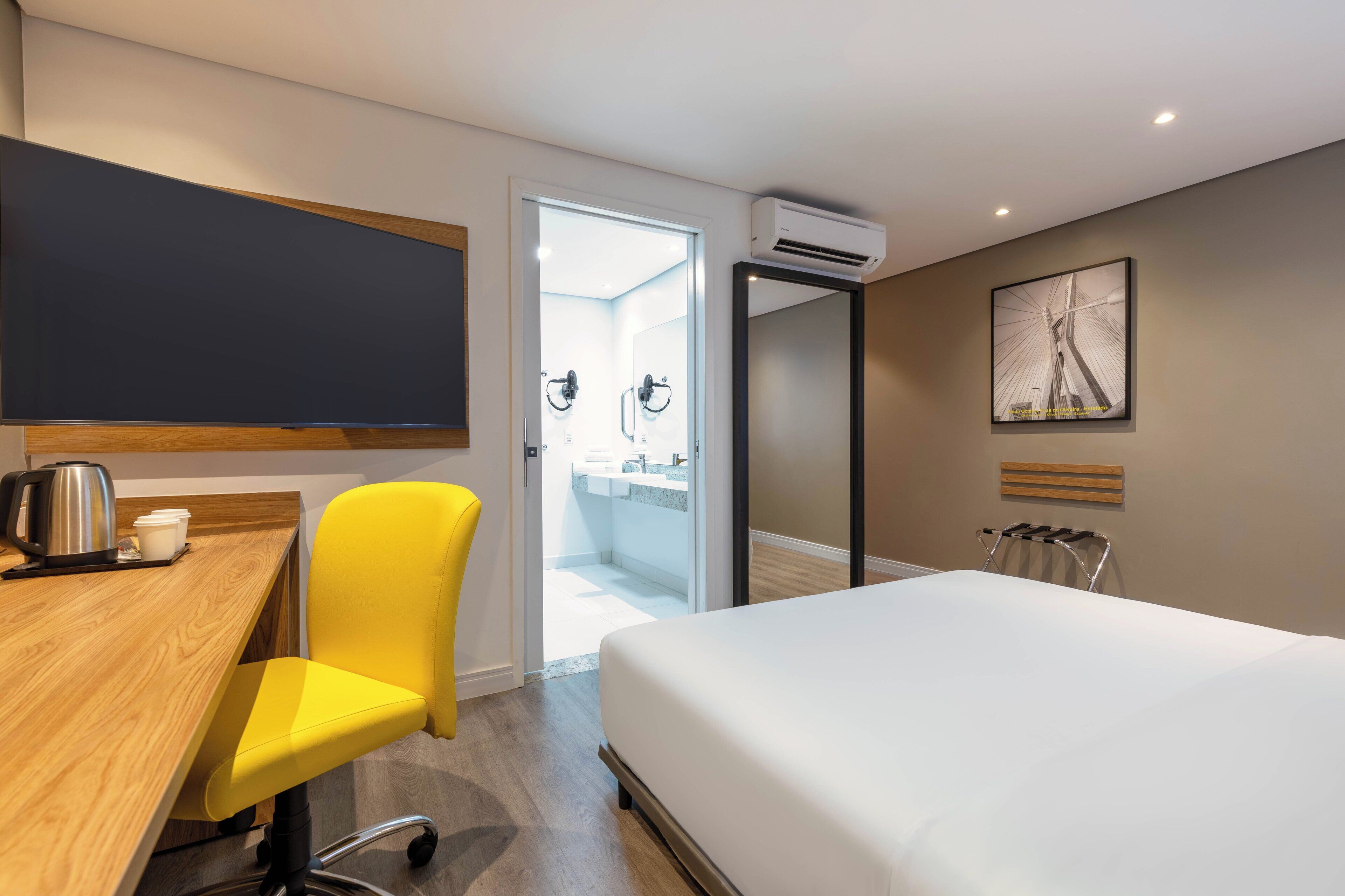Habitación de hotel con cama, escritorio y silla, cafetera, aire acondicionado, baño con ducha y lavamanos, y un cuadro en la pared.