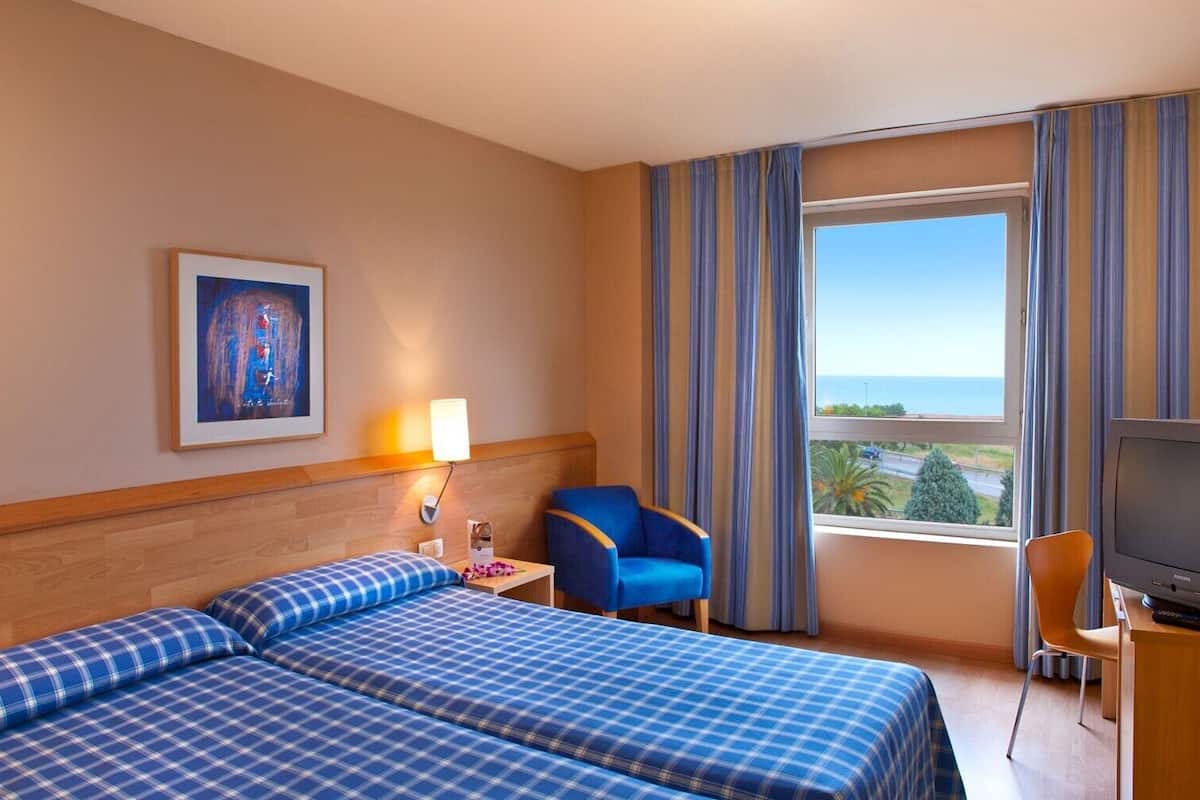 Una habitación de hotel con dos camas, un escritorio con televisor, una silla, una lámpara y una ventana con cortinas.